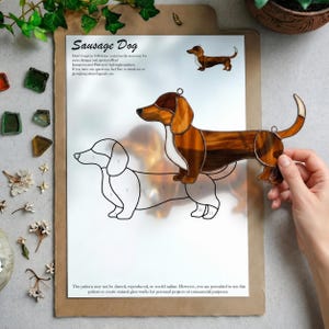 May include: A stained glass dachshund suncatcher featuring brown and white glass pieces held together by dark metal solder lines. The piece is held over a printed paper pattern sheet titled Sausage Dog that outlines the numbered glass segments.