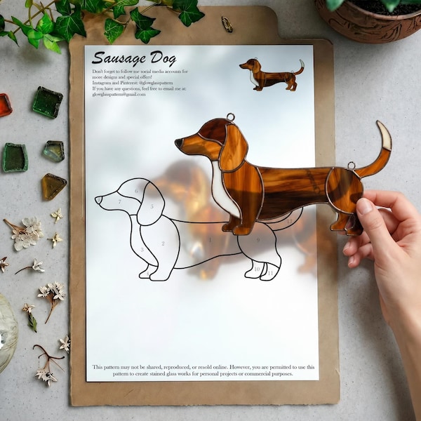 Puede incluir: Un atrapasoles de vitral con forma de perro salchicha, hecho con piezas de vidrio marrón y blanco unidas por líneas de soldadura metálica oscura. La pieza se sostiene sobre una hoja de patrón de papel titulada Sausage Dog con segmentos.