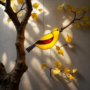 May include: A stained glass bird ornament with yellow and red glass. The bird is hanging from a string in front of a tree branch with yellow leaves.
