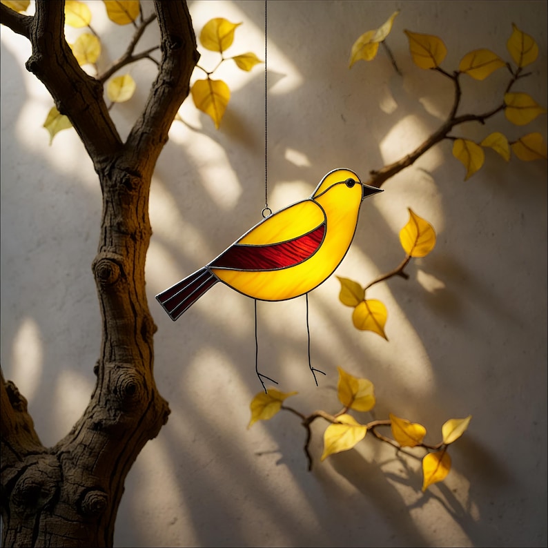 May include: A stained glass bird ornament with yellow and red glass. The bird is hanging from a string in front of a tree branch with yellow leaves.