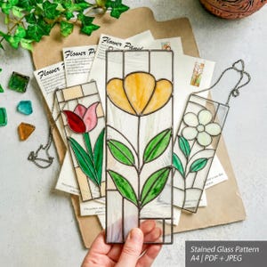May include: Three rectangular stained glass panels featuring floral designs. One panel shows a yellow flower, another a red tulip, and the third a white flower. Each panel has a metal frame and a hanging chain. Text on the paper reads Stained Glass Pattern A4 PDF + JPEG.