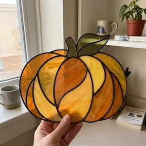 May include: A stained glass pumpkin suncatcher featuring various shades of orange and yellow glass segments with a dark green glass leaf at the top. The individual glass pieces are joined together by dark metal solder lines in a curved pattern.
