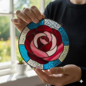 May include: A round stained glass piece featuring a rose design. The rose is depicted with shades of red, pink, and white, surrounded by blue and silver-colored glass. The piece is held up by a person.