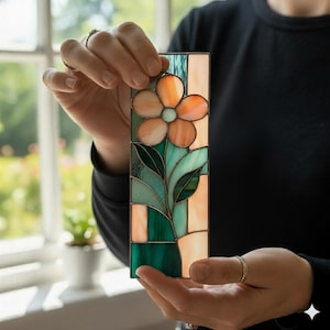 May include: A rectangular stained glass panel featuring a central peach-colored flower with five petals and a light green center. The design includes dark green leaves and stems set against a background of vertical peach and teal glass segments.