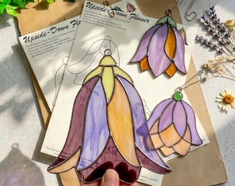 Könnte beinhalten: Drei Blumen-Ornamente aus Buntglas in Lila- und Gelbtönen mit Metall-Lötlinien. Zwei fertige Stücke hängen an Ketten, während ein größeres Stück über einer Papiervorlage mit der Aufschrift Upside-Down Flower in der Hand gehalten wird.