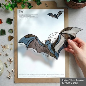 May include: A stained glass bat pattern on an A4 sheet of paper, featuring a finished bat-shaped stained glass piece with brown and blue glass segments. The paper includes the text Bat, social media handles, and usage permissions for the pattern.