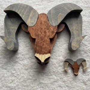May include: Wooden ram head wall art in shades of brown and gray. The ram's head is made of wood and features detailed horns and a textured finish. A smaller version of the ram head is also present. The artwork is likely for home decor.
