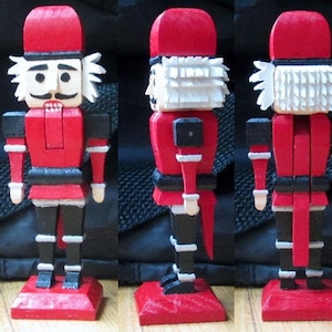 May include: A red and black wooden nutcracker with a white beard and a red hat. The nutcracker is standing on a red base.