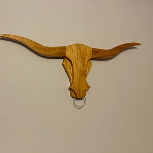 May include: A wooden wall decor in the shape of a longhorn steer head with a metal ring hanging from the bottom.