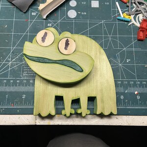 Puede incluir: Un proyecto artesanal en forma de rana de madera con un acabado verde claro. La rana tiene ojos grandes y redondos con pupilas marrones y una boca verde oscuro. La rana mide unos 15 cm de largo.