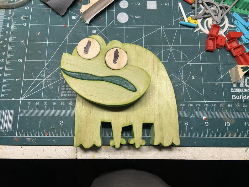 Puede incluir: Un proyecto artesanal en forma de rana de madera con un acabado verde claro. La rana tiene ojos grandes y redondos con pupilas marrones y una boca verde oscuro. La rana mide unos 15 cm de largo.