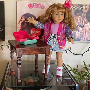 May include: A doll wearing a pink and purple cardigan over a pink shirt and a denim skirt. The doll is standing next to a small wooden table with a red and green toy on top. The table has four legs with decorative carvings. The doll is wearing white sneakers. The background includes a record album cover with the text "Dreamboat Annie" and a string of Christmas lights.