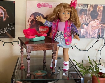 Può includere: Una bambola che indossa un cardigan rosa e viola sopra una camicia rosa e una gonna di jeans. La bambola è in piedi accanto a un piccolo tavolo di legno con un giocattolo rosso e verde sopra. Il tavolo ha quattro gambe con intagli decorativi. La bambola indossa scarpe da ginnastica bianche. Lo sfondo include una copertina di un album discografico con il testo "Dreamboat Annie" e una ghirlanda di luci natalizie.