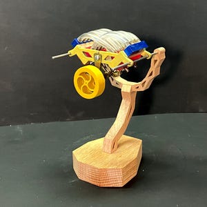 Könnte beinhalten: Eine Holzskulptur eines mechanischen Geräts mit einem gelben Rad und einer transparenten, gebogenen Oberseite. Das Gerät wird von einem Holzständer mit sechseckiger Basis getragen. Die Skulptur hat gelbe, blaue und rote Akzente.