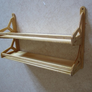 Può includere: Una mensola in legno chiaro a due livelli, montata a parete. La mensola ha un design semplice con due piattaforme orizzontali e supporti laterali decorativi. Il legno sembra non rifinito o leggermente tinto.