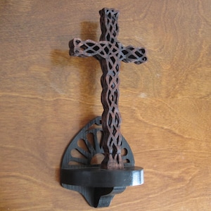 May include: A wooden cross with a decorative, intricate design. The cross is mounted on a black wooden wall mount with a decorative design.