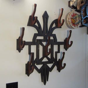 Pode incluir: Um cabide de parede decorativo de madeira escura com oito ganchos castanhos. O design apresenta formas geométricas e um elemento central pontiagudo, criando uma solução de armazenamento única e artística.