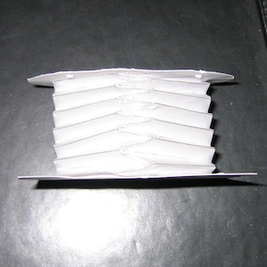May include: A white ribbon or trim is neatly wound around a flat, rectangular cardboard spool. The material is folded in a repeating zigzag pattern, creating a layered, accordion-like appearance between the top and bottom edges of the spool.