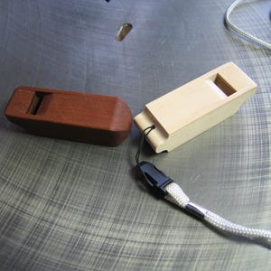 May include: Two wooden whistles, one dark brown and one light beige, are displayed. The brown whistle has a rectangular shape, while the beige whistle has a rectangular shape with a lanyard attached. The whistles are on a metallic surface.