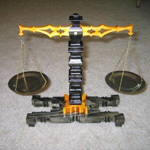 May include: A decorative balance scale with a black and gold color scheme. The scale features two oval-shaped pans suspended by gold chains from a decorative arm. The base and central column are black, with gold accents.