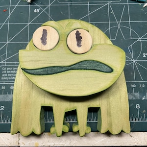 Könnte beinhalten: Eine handgefertigte Holzfroschskulptur mit hellgrüner Oberfläche. Der Frosch hat große, runde Augen mit dunkelbraunen Pupillen und einen grünen Mund. Die Beine des Frosches sind ausgeschnitten. Auf einer grünen Schneidematte.