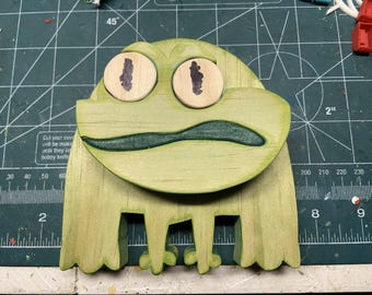 Peut inclure: Sculpture de grenouille en bois peint en vert clair. La grenouille a de grands yeux ronds avec des pupilles marron foncé et une bouche verte. Les pattes de la grenouille sont découpées. Sur un tapis de coupe vert.