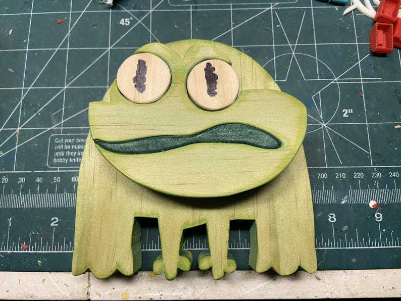 Puede incluir: Escultura de rana de madera pintada de verde claro. La rana tiene ojos grandes y redondos con pupilas marrones oscuras y una boca verde. Las patas de la rana est&aacute;n recortadas. Sobre una esterilla de corte verde.