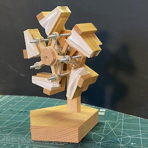 May include: A wooden kinetic sculpture with a wheel-like design and multiple blades. The sculpture is made from light-coloured wood, with silver hardware and white accents. It stands on a rectangular wooden base.