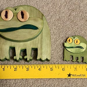 Puede incluir: Dos figuras de rana de madera. La rana m&aacute;s grande mide aproximadamente 10 cm de ancho y 10 cm de alto, con cuerpo verde, ojos marrones y boca verde oscuro. La rana m&aacute;s peque&ntilde;a mide unos 5 cm de ancho y 5 cm de alto. Una regla amarilla en la parte inferior.