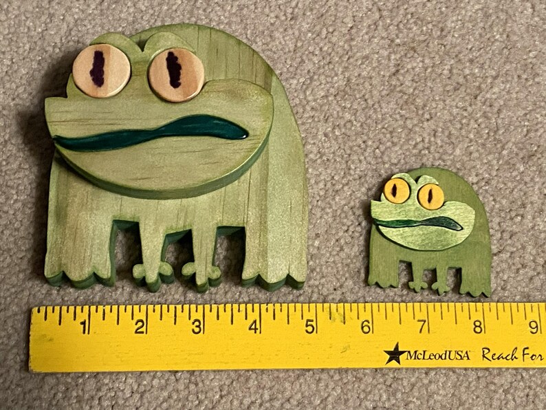 Puede incluir: Dos figuras de rana de madera. La rana m&aacute;s grande mide aproximadamente 10 cm de ancho y 10 cm de alto, con cuerpo verde, ojos marrones y boca verde oscuro. La rana m&aacute;s peque&ntilde;a mide unos 5 cm de ancho y 5 cm de alto. Una regla amarilla en la parte inferior.