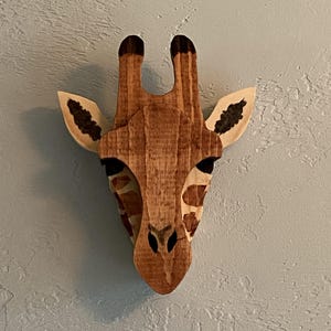 May include: Wooden giraffe head wall decor. The giraffe head is made of stained wood with a natural wood grain finish. The giraffe has dark brown spots and black accents on the ears and horns. The giraffe is mounted on a light blue wall.