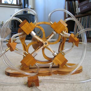 Könnte beinhalten: Eine komplexe Skulptur aus Holz und transparenten Schläuchen. Sie zeigt ein zentrales Holzrad mit radialen Armen, verbunden mit Holzblöcken und transparenten Schläuchen. Die Basis ist ebenfalls aus Holz gefertigt.