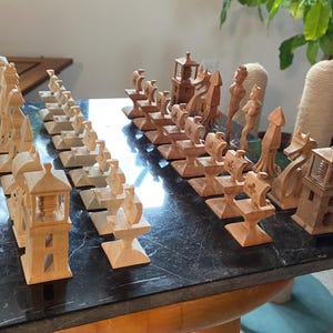 Puede incluir: Un juego de ajedrez de madera con piezas talladas intrincadamente. Las piezas incluyen torres, figuras y otras formas tradicionales de ajedrez. El juego se exhibe sobre una superficie oscura, similar al mármol.