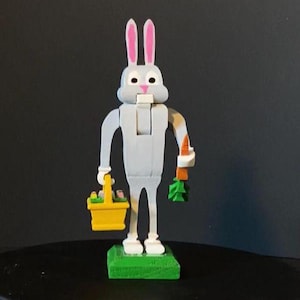 Puede incluir: Una figurita de conejo gris con orejas rosas, sosteniendo una zanahoria y una cesta amarilla. El conejo está de pie sobre una base verde.