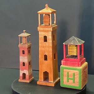 May include: Three wooden toy lighthouses, one red and white, one brown, and one with a green block base with the letter "H" on it.