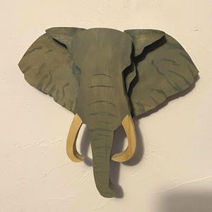 Op de afbeelding: Houten sculptuur van een olifantenkop met grote oren en slagtanden. De olifant is geschilderd in grijstinten en groen, met lichtbruine slagtanden. De sculptuur is aan een witte muur bevestigd.