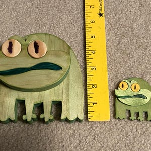 Puede incluir: Dos figuras de rana de madera en tonos verdes, con ojos amarillos y marrones y boca verde oscuro. Una regla amarilla se coloca junto a las ranas. La rana m&aacute;s grande mide aproximadamente 13 cm de largo.