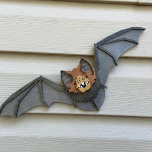 May include: A layered wooden wall decoration shaped like a bat with outstretched wings. The bat features a dark grey wing structure, a textured brown fur body, and a light tan face with pointed ears and a mouth showing sharp white teeth.