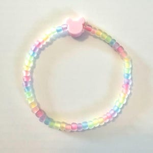 May include: A beaded bracelet featuring a rainbow of pastel colors. The bracelet is made of small, round beads and has a pink bear-shaped clasp. The beads transition from yellow to pink, blue, and green.