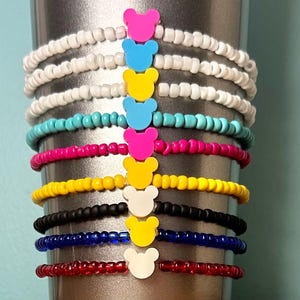 May include: A collection of beaded bracelets in various colors, including white, blue, pink, yellow, black, and red. Each bracelet features a colorful bear-shaped charm. The bracelets are stacked together, showcasing a variety of vibrant hues.