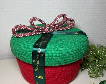 Puede incluir: Una cesta de Navidad redonda y tejida, con base roja y tapa verde. Una cinta verde con texto "Merry Christmas" envuelve la cesta. Un lazo de cuerda rojo y blanco encima. Tema navideño festivo.