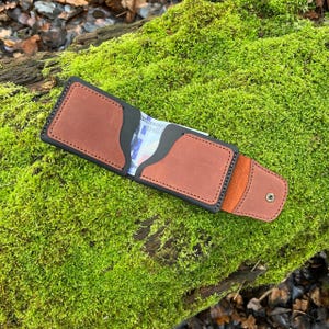 May include: A brown leather wallet with black edges and a snap closure. The wallet is open, revealing a section for cash and cards. The wallet is resting on a bed of green moss.