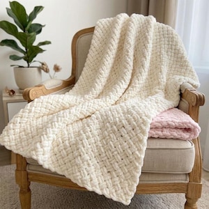 May include: A cream-colored, chunky knit blanket draped over a vintage-style chair. The blanket has a woven texture and is folded over a smaller, pink blanket. The chair has a light wood frame and a beige cushion. A potted plant is visible in the background.