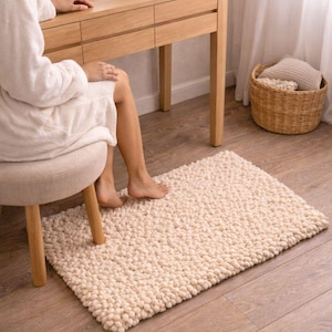 May include: A cream-colored, rectangular bath mat with a textured, nubby surface. The rug is placed on a wooden floor near a vanity and a small stool. A wicker basket and a window with sheer curtains are in the background.