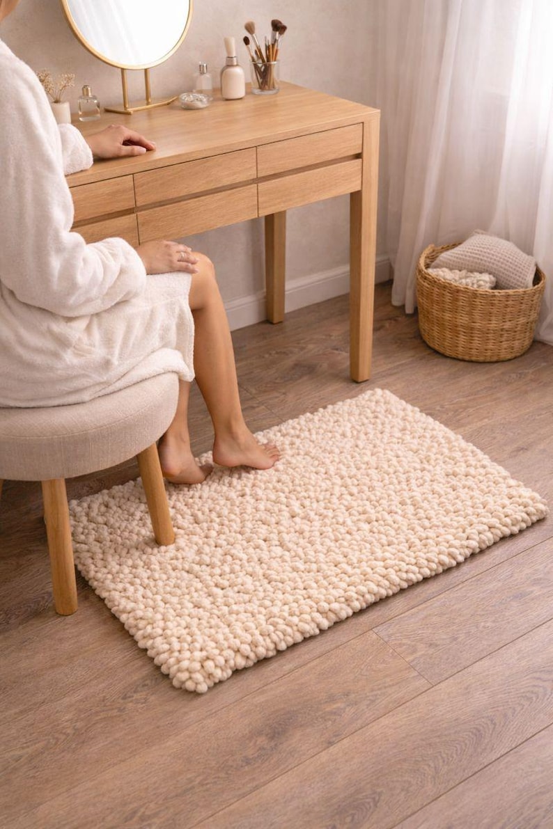 May include: A cream-colored, rectangular bath mat with a textured, nubby surface. The rug is placed on a wooden floor near a vanity and a small stool. A wicker basket and a window with sheer curtains are in the background.