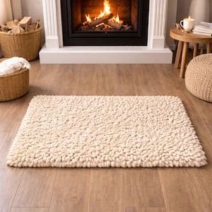 May include: A rectangular, cream-colored rug with a textured, nubby surface. The rug is placed on a hardwood floor in front of a fireplace with a burning fire. A white mantel and decorative items are in the background.