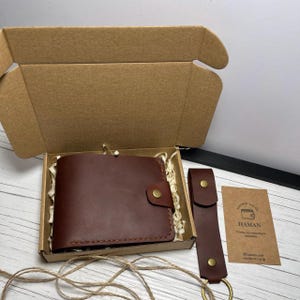 May include: A brown leather wallet with a snap closure and a matching key fob are presented in a cardboard box with packing material. A small card with text is also included.