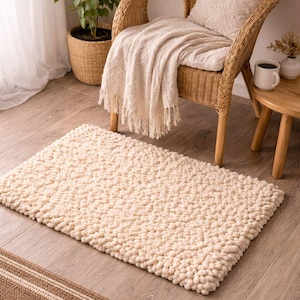 May include: A cream-colored, textured rug with a dense, looped pile. The rug is rectangular and sits on a wooden floor. A woven wicker chair with a cream-colored throw and pillow is in the background, along with a small side table.
