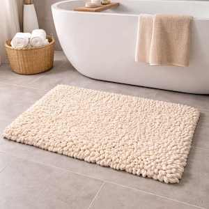 May include: A rectangular, cream-colored bath mat with a textured, looped surface. The mat is placed on a gray tiled floor in front of a white bathtub. A woven basket with rolled white towels and a vase with dried plants are also visible.