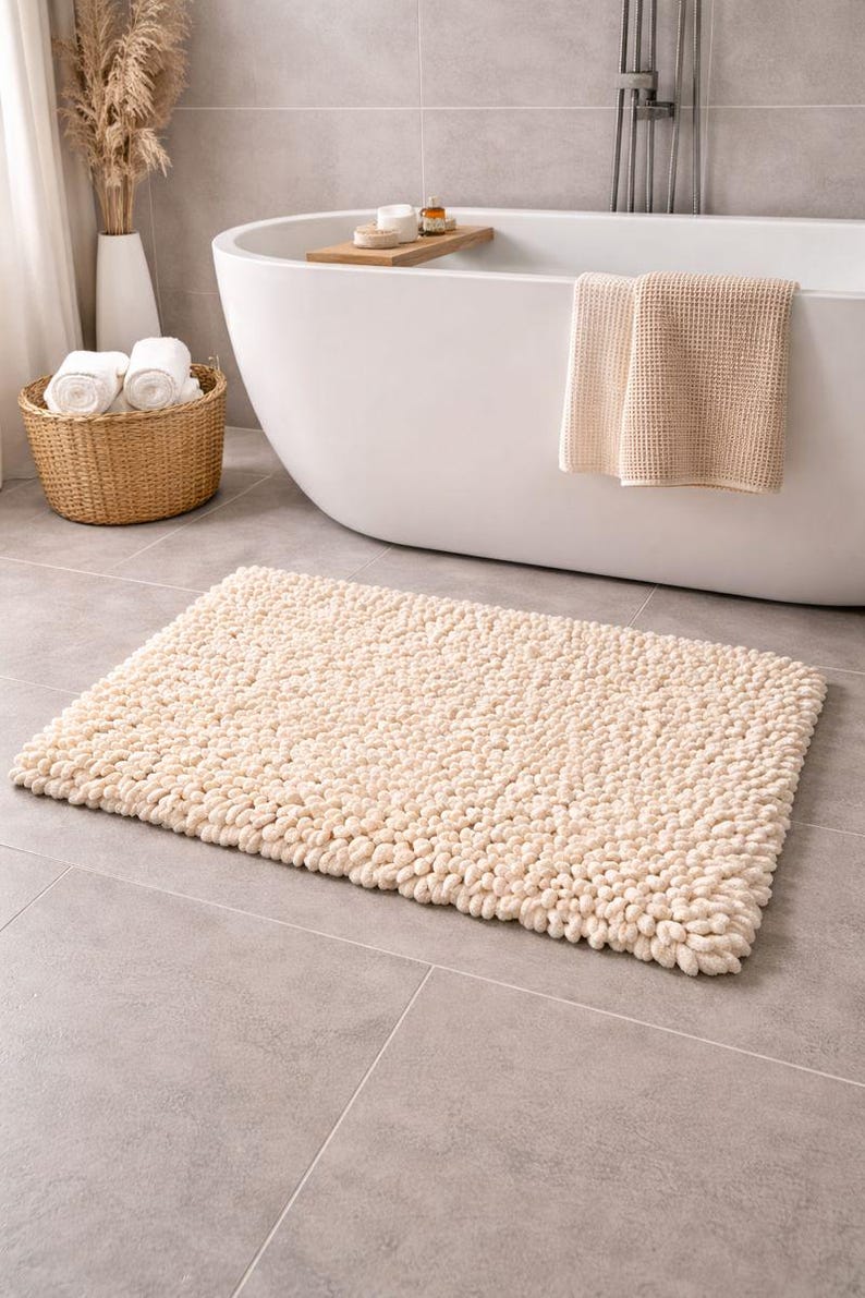 May include: A rectangular, cream-colored bath mat with a textured, looped surface. The mat is placed on a gray tiled floor in front of a white bathtub. A woven basket with rolled white towels and a vase with dried plants are also visible.